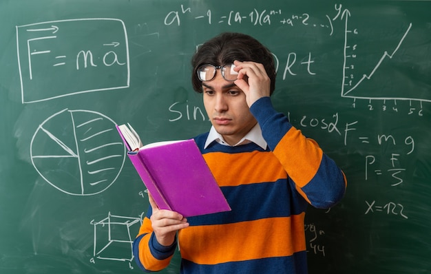 Concentrated young caucasian geometry teacher wearing glasses standing in front of chalkboard in classroom raising glasses reading book