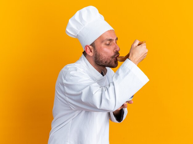 Concentrated young caucasian male cook in chef uniform and cap standing in profile view holding bowl testing meal with spoon isolated on orange wall