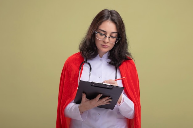 Concentrated young caucasian superhero girl in red cape wearing doctor uniform and stethoscope with glasses writing on clipboard with pencil 