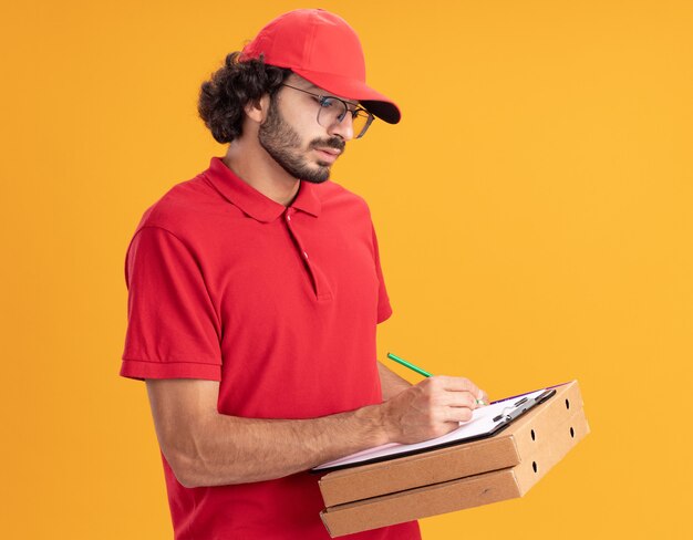 Сосредоточенный молодой доставщик в красной форме и кепке в очках, стоящий в профиль, держа пакеты с пиццей, пишущие в буфере обмена с карандашом, изолированным на оранжевой стене