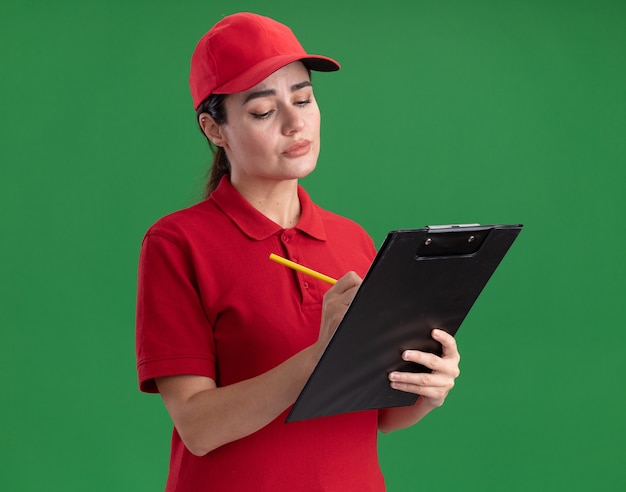 Concentrated young delivery woman in uniform and cap writing on clipboard with pencil 