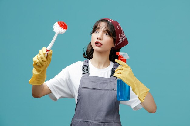 균일한 반다나와 고무 장갑을 끼고 브러시를 들고 측면을 바라보는 세척제를 착용한 집중된 젊은 여성 청소부는 파란색 배경에 고립된 것을 청소하는 척