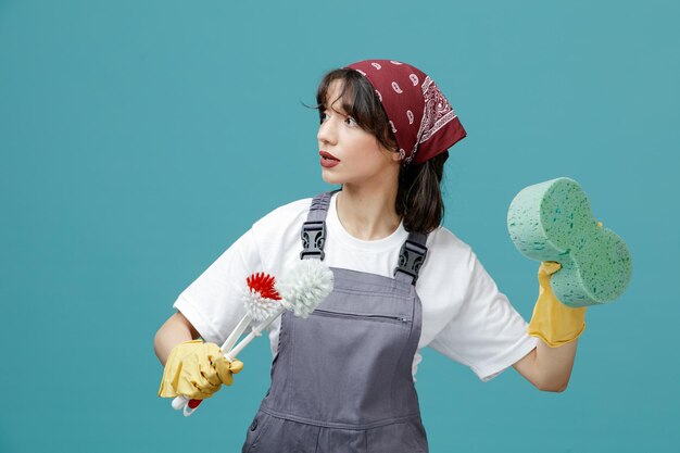 Концентрированная молодая женщина-уборщица в униформе бандана и резиновых перчатках, держащая туалетные щетки и губку в воздухе и смотрящая в сторону, изолированную на синем фоне