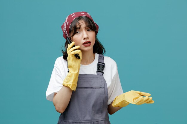 Concentrated young female cleaner wearing uniform bandana and rubber gloves talking on phone looking at side showing empty hand isolated on blue background