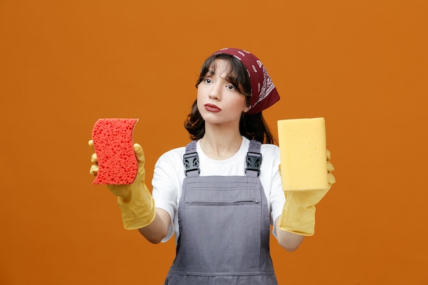 Concentrated young female cleaner wearing uniform rubber gloves and bandana holding sponges looking at side pretend cleaning something isolated on orange background