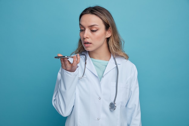 Concentrated young female doctor wearing medical robe and stethoscope around neck holding and looking at mobile phone talking by its microphone 