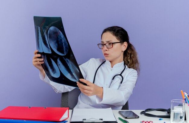 Concentrated young female doctor wearing medical robe and stethoscope sitting at desk with medical tools holding and looking at x-ray shot isolated