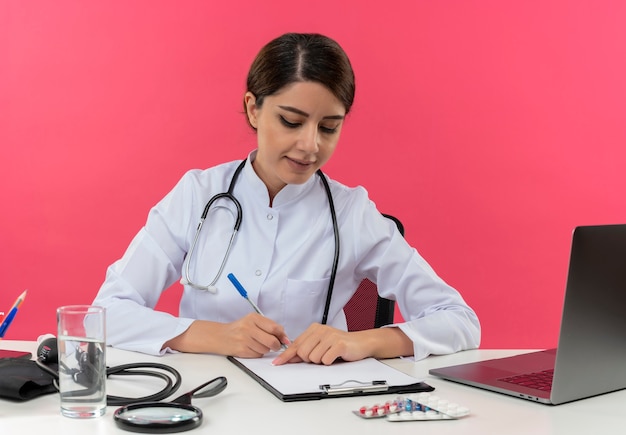 Concentrated young female doctor wearing medical robe and stethoscope sitting at desk with medical tools and laptop writing with pen on clipboard isolated on pink wall