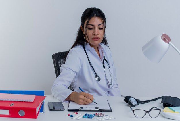 Concentrated young female doctor wearing medical robe and stethoscope sitting at desk with medical tools writing on clipboard isolated