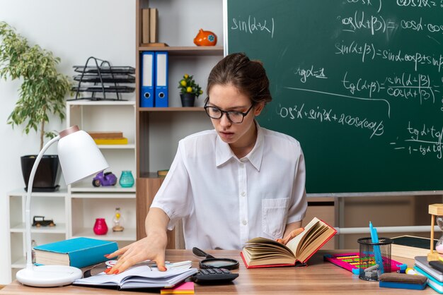 concentrated young female math teacher wearing glasses sitting at desk with school supplies grabbing open book keeping hand on open note pad looking down at magnifying glass in classroom