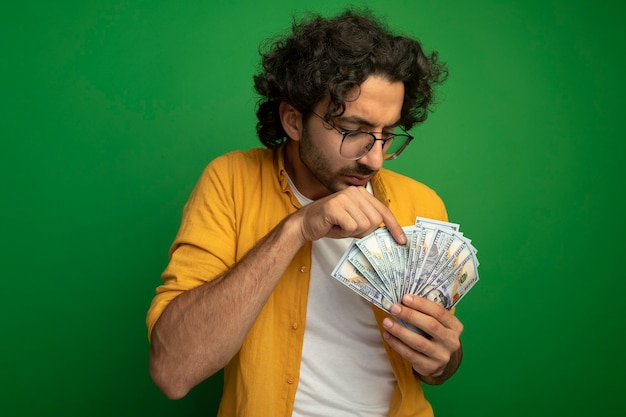 Concentrato giovane uomo caucasico bello con gli occhiali che contano soldi isolati sulla parete verde con lo spazio della copia