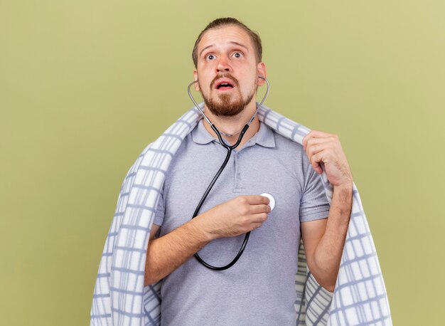 Concentrated young handsome slavic ill man wearing stethoscope listening to his own heartbeat wrapped in plaid grabbing plaid looking up isolated on olive green wall with copy space