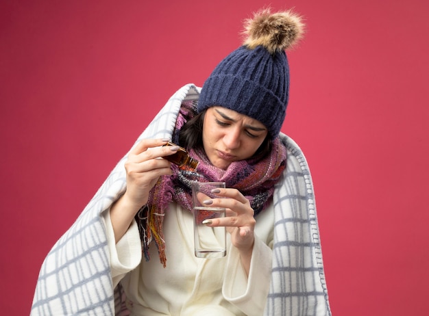 Сосредоточенная молодая больная женщина в халате, зимней шапке и шарфе, завернутом в плед, добавляя лекарство в стакан с водой, изолированный на розовой стене