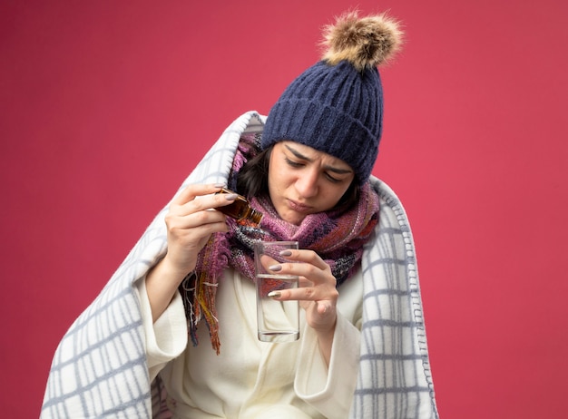 Concentrated young ill woman wearing robe winter hat and scarf wrapped in plaid adding medicament into glass of water isolated on pink wall