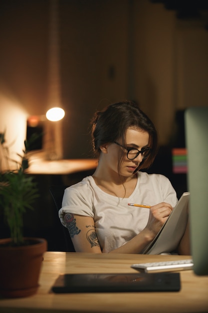 Concentrated young lady designer sitting indoors at night writing notes.