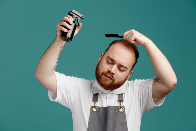 Concentrated young male barber wearing white shirt and barber apron combing his hair applying hair spray on his own hair with closed eyes isolated on blue background