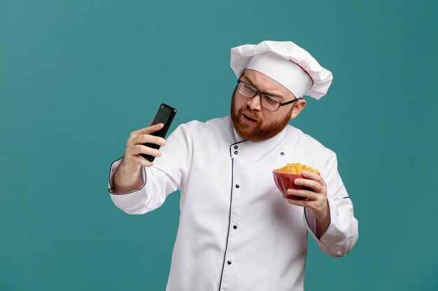 Concentrated young male chef wearing glasses uniform and cap holding bowl of macaronis and mobile phone taking selfie isolated on blue background