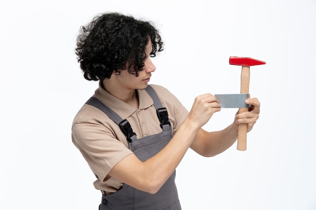 Concentrated young male construction worker wearing uniform taping hammer with scotch tape isolated on white background