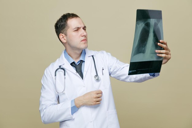 Concentrated young male doctor wearing medical robe and stethoscope around neck holding xray shot looking at it isolated on olive green background