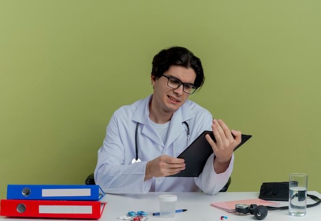 Free photo concentrated young male doctor wearing medical robe and stethoscope with glasses sitting at desk with medical tools holding and looking at clipboard isolated