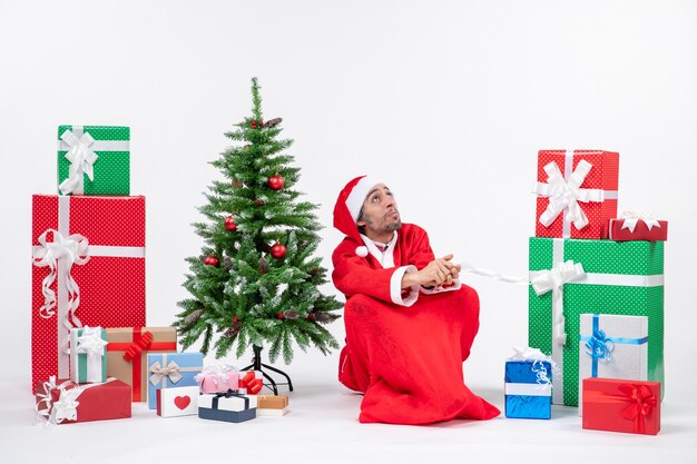 Concentrated young man dressed as Santa claus with gifts and decorated Christmas tree sitting on the ground on white background