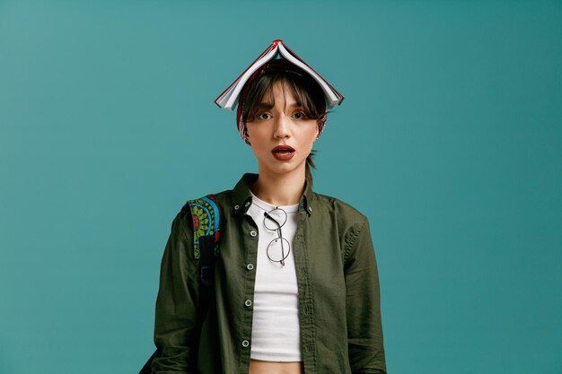 Concentrated young student girl wearing bandana and backpack putting glasses on her blouse looking at camera holding open note pad on her head isolated on blue background