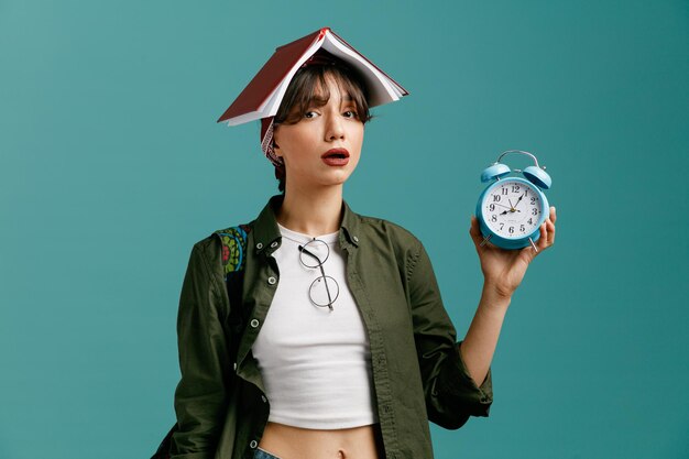 Concentrated young student girl wearing bandana and backpack putting glasses on her blouse showing alarm clock looking at camera holding open note pad on her head isolated on blue background