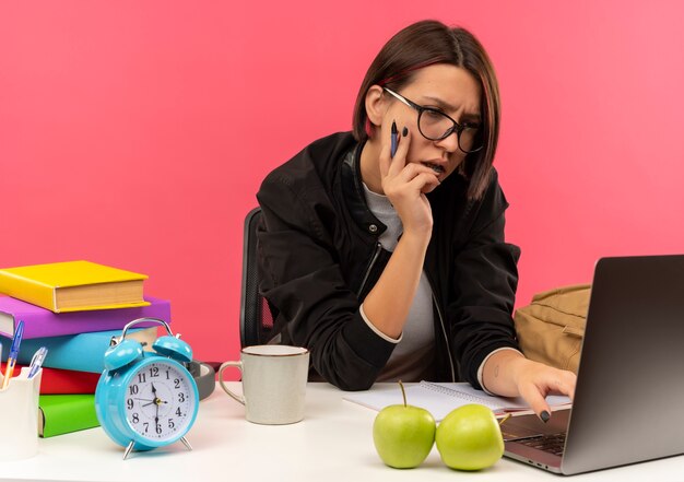 Концентрированная молодая студентка в очках сидит за столом, держа ручку, кладя руку на лицо, используя ноутбук, изолированный на розовом