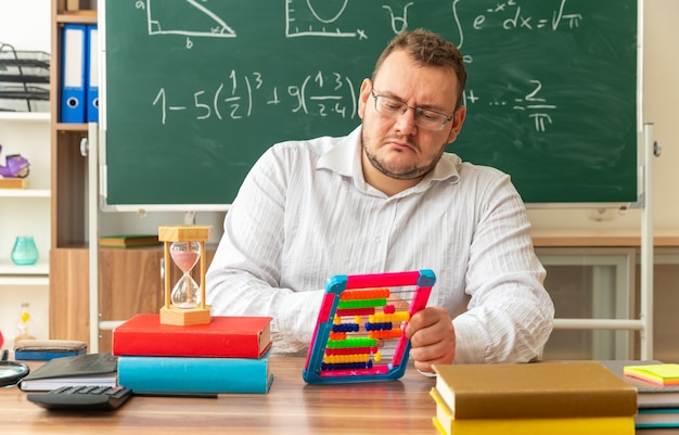 concentrated young teacher wearing glasses sitting at desk with school supplies in classroom using abacus