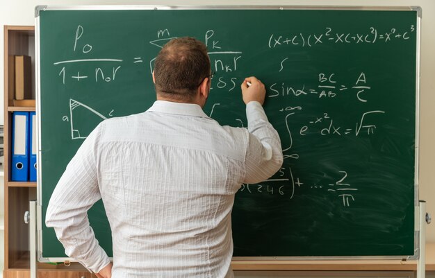 concentrated young teacher wearing glasses standing in behind view in front of chalkboard in classroom keeping hand on waist writing formula on chalkboard with chalk