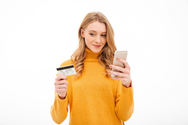 Сконцентрированная молодая женщина держа мобильный телефон и кредитную карточку.