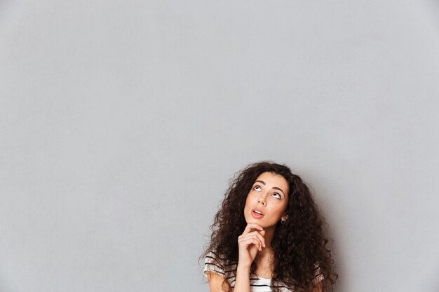 Concentrated young woman with shaggy hair touching her chin with face upward and thinking or dreaming over grey wall 