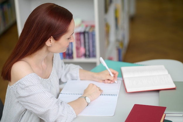 Концентрация. серьезная молодая женщина с длинными рыжими волосами в профиле перед камерой сидит и пристально пишет за столом с книгами и ноутбуком в помещении