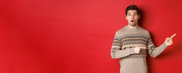 Concept of christmas celebration winter holidays and lifestyle Amazed handsome guy in xmas sweater pointing fingers right at logo showing advertisement standing over red background