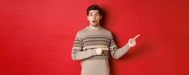 Concept of christmas celebration, winter holidays and lifestyle. Amazed handsome guy in xmas sweater pointing fingers right at logo, showing advertisement, standing over red background.