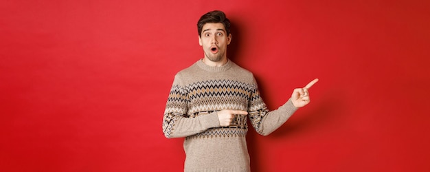 Concept of christmas celebration, winter holidays and lifestyle. Amazed handsome guy in xmas sweater pointing fingers right at logo, showing advertisement, standing over red background.