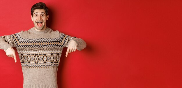 Concept of christmas celebration, winter holidays and lifestyle. Excited caucasian male model in xmas sweater, pointing fingers down and making announcement, showing advertisement, red background