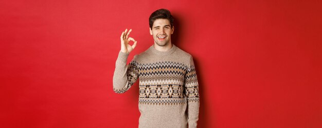 크리스마스 축 하, 겨울 휴가 및 라이프 스타일의 개념입니다. 크리스마스 스웨터를 입은 잘 생기고 자신감 있는 남자의 이미지, 무언가를 보장하고, 괜찮은 표시와 미소, 빨간색 배경을 보여줍니다.