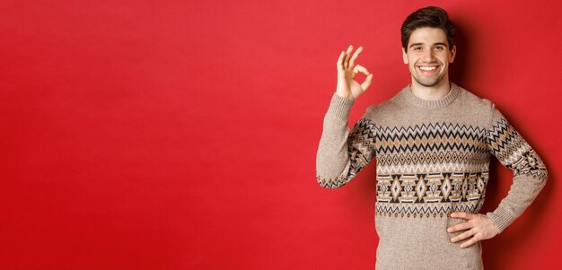 Concept of christmas celebration winter holidays and lifestyle Portrait of attractive man in xmas sweater smiling happy and showing okay sign praise something good red background