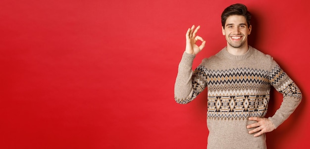 Концепция празднования Рождества, зимних праздников и образа жизни Портрет привлекательного мужчины в рождественском свитере, счастливо улыбающегося и показывающего хороший знак