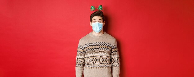 Концепция рождественского ковида и социального дистанцирования веселый молодой человек в медицинской маске и новогодней одежде
