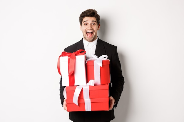 Concept of christmas holidays, celebration and lifestyle. Image of handsome amazed guy in suit, holding new year presents and smiling, standing against white background