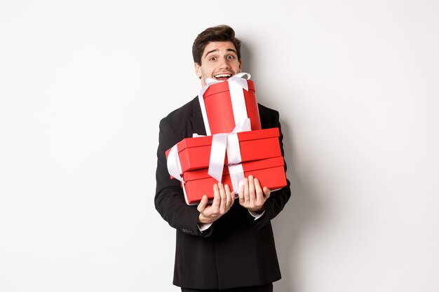 クリスマス休暇、お祝い、ライフスタイルの概念。スーツを着た幸せな男の画像は、新年のプレゼントを運び、贈り物と笑顔で箱を持って、白い背景に立って