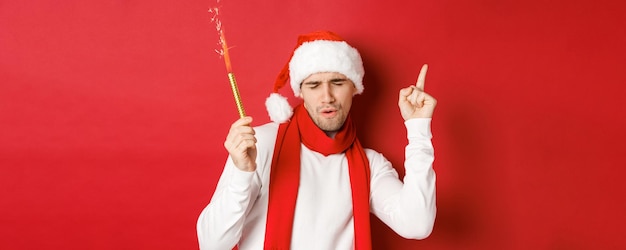 クリスマス、冬休み、お祝いのコンセプトです。新年会を楽しんだり、線香花火で踊ったり、サンタの帽子とスカーフを身に着けたり、赤い背景の上に立ったりする魅力的な男。