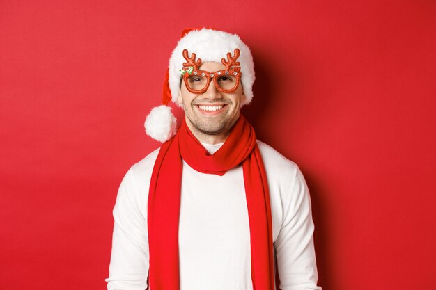 Concept of christmas, winter holidays and celebration. Cheerful handsome guy in new year party glasses and smiling, standing over red background