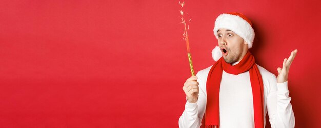 Концепция Рождества, зимних праздников и празднования. Крупный план изумленного красивого мужчины в новогодней шапке и шарфе, с волнением смотрящего на бенгальский огонь, стоящего на красном фоне.