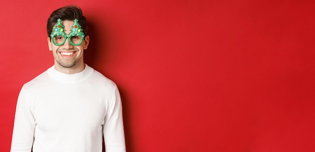 Free photo concept of christmas, winter holidays and celebration. close-up of handsome man in white sweater and party glasses, enjoying new year, standing over red background.