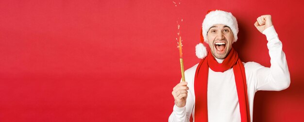 Concept of christmas winter holidays and celebration Handsome man celebrating new year and having fun holding sparkler and smiling wearing santa hat standing over red background