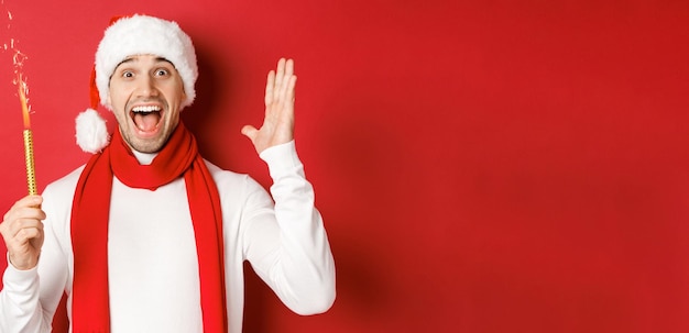 Концепция Рождества, зимних праздников и празднования. Красивый мужчина празднует Новый год и веселится, держит бенгальский огонь и улыбается, в шляпе санта-клауса, стоя на красном фоне.