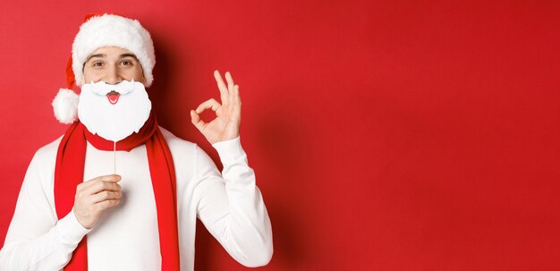 Concept of christmas winter holidays and celebration Pleased handsome man in santa hat holding long white beard mask and showing okay sign standing over red background